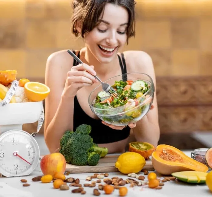 Smiling woman eating a fresh salad at a kitchen table with fruit and vegetables